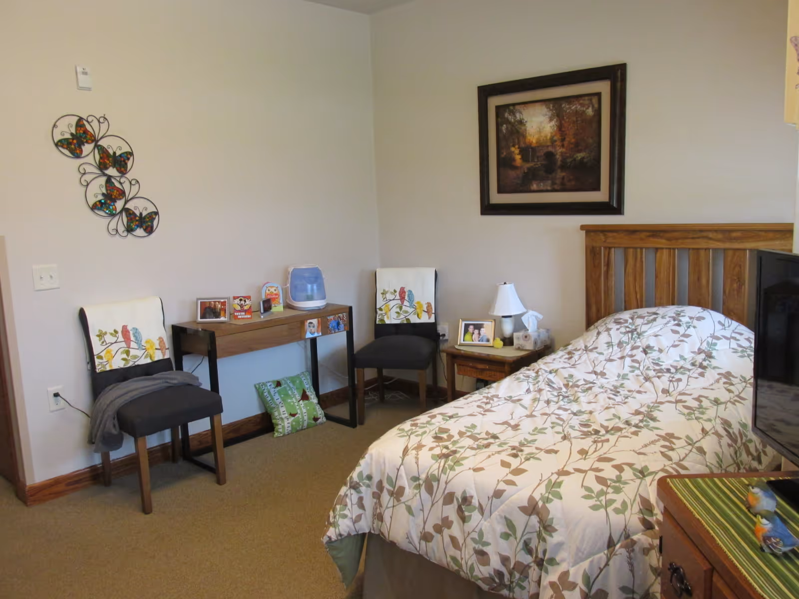 A cozy bedroom with a wooden bed frame and a floral patterned comforter. There are two chairs with bird-themed cushions, a small wooden desk with personal items and photos, and a nightstand with a lamp and framed photo. A framed landscape painting hangs on the wall above the bed, and decorative butterfly wall art is on the adjacent wall.