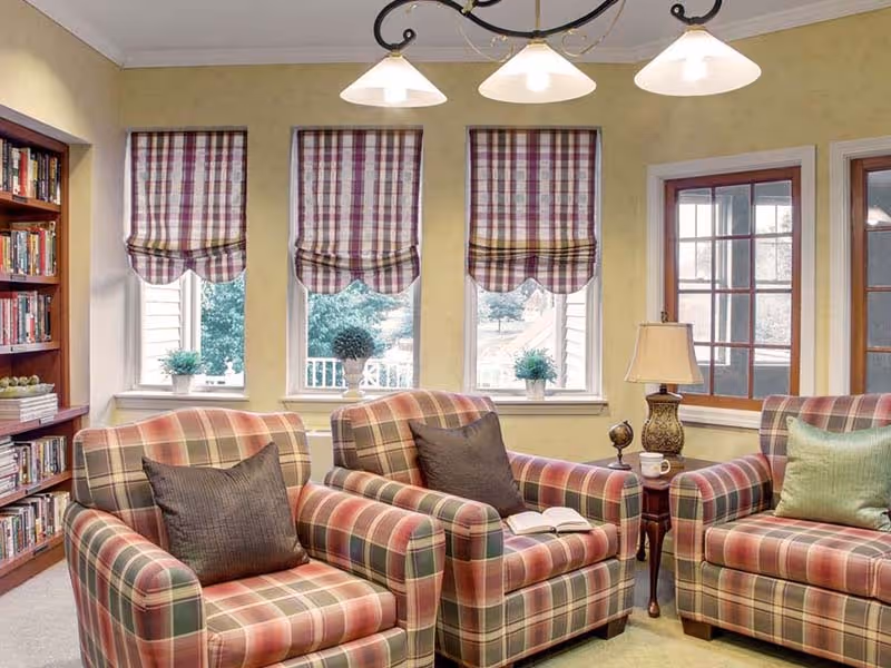 A cozy living room area with three plaid upholstered armchairs arranged around a small wooden side table holding a lamp and a small decorative globe. Behind the chairs are three windows with plaid Roman shades and small potted plants on the windowsills. To the left is a wooden bookshelf filled with books.