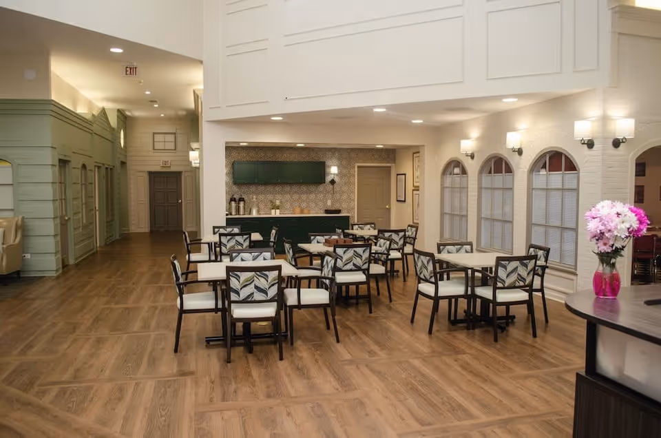 A bright and spacious dining area with several square tables and chairs featuring patterned cushions. The room has wood flooring, light-colored walls, and a small counter area with cabinets and coffee dispensers in the background. There are arched windows with blinds on one side and a vase with pink and white flowers on a round table in the foreground.