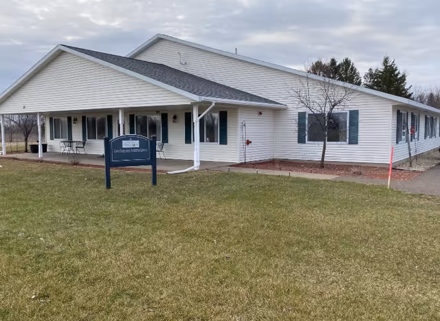 Single-story white senior living building with a covered front porch, a lawn sign, and green shutters.