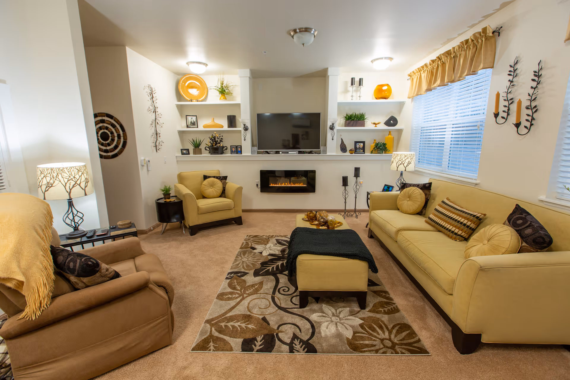 Bright, warmly decorated living room with beige sofas and armchairs around a patterned rug, a TV above a wall fireplace, built-in shelves, and lamps.