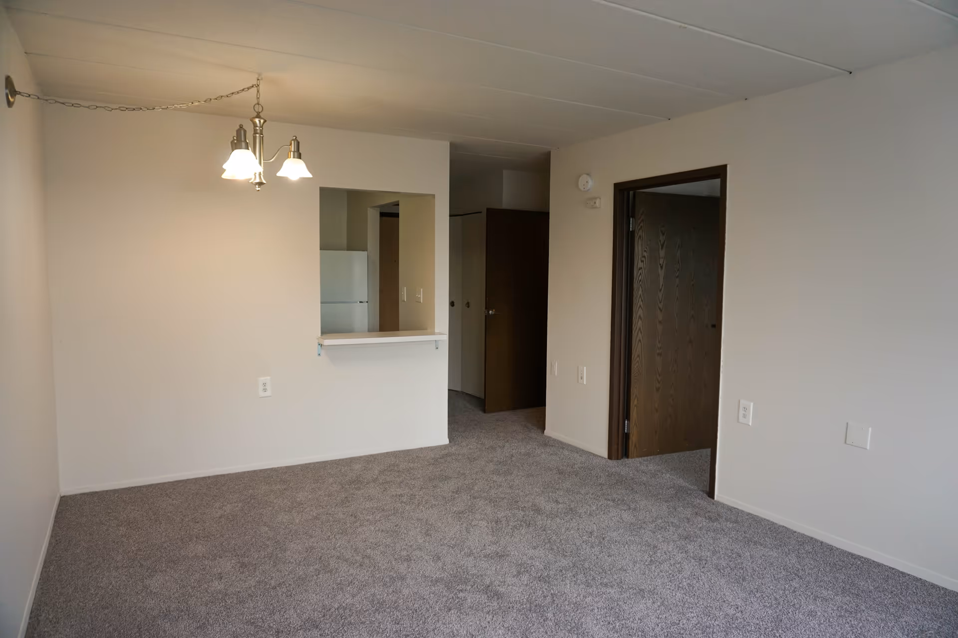 Empty room with beige walls and gray carpet, featuring a hanging light fixture, a pass-through window to a kitchen area with a visible refrigerator, and an open doorway leading to another room.