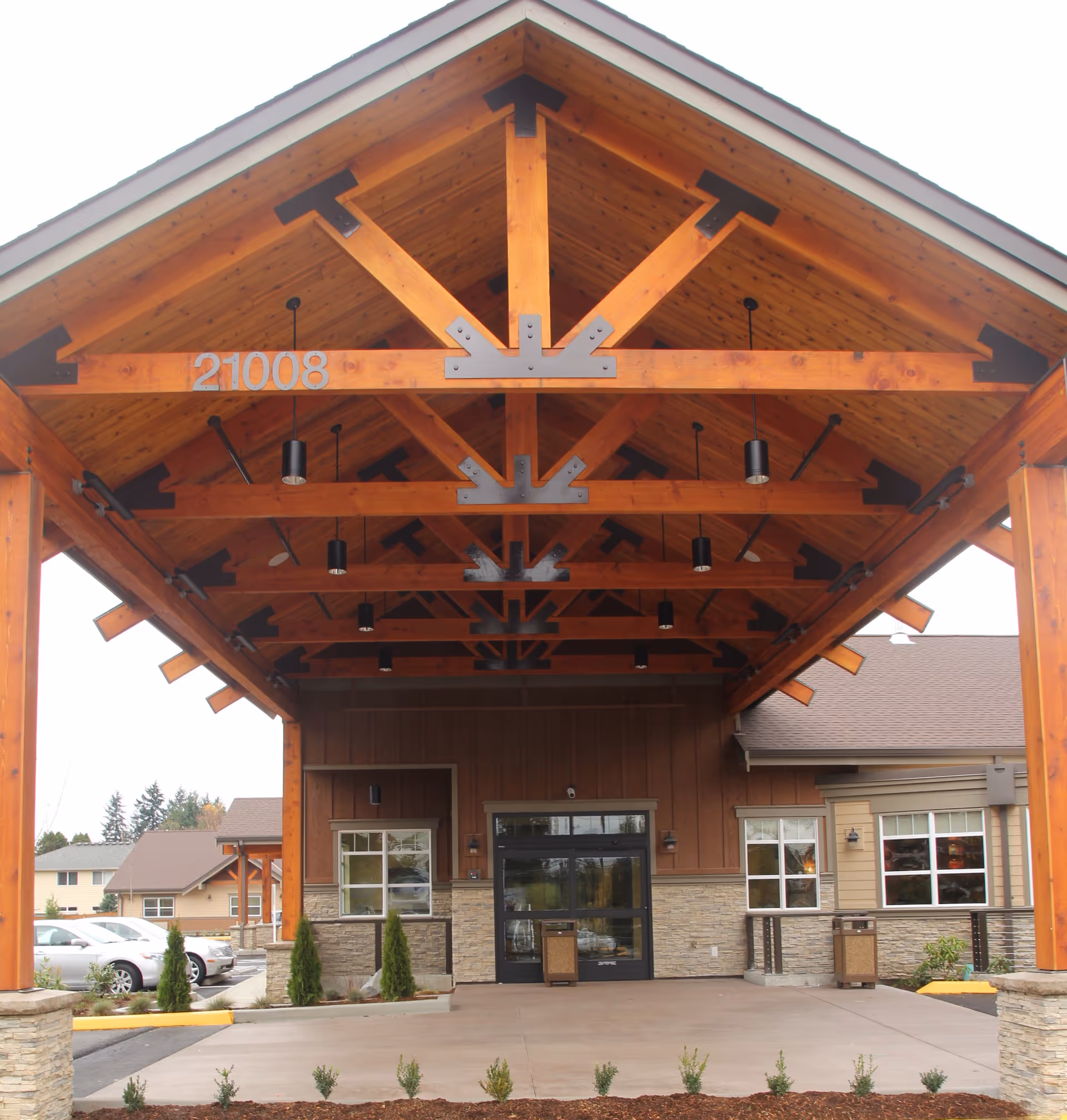 Entrance of a building with a large wooden canopy supported by wooden beams and metal brackets. The building has stone and wood siding, with windows on either side of a glass door entrance. Several small shrubs are planted in front, and cars are parked to the left side.