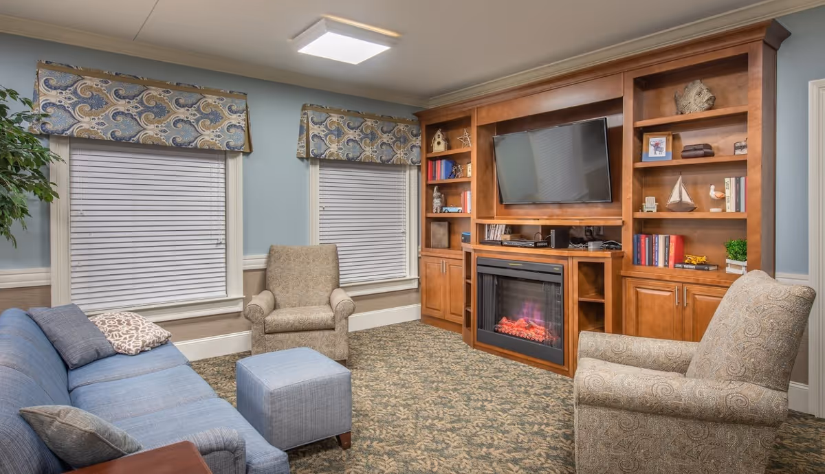 A cozy living room with a blue sofa, two patterned armchairs, and a blue ottoman. The room features a built-in wooden entertainment center with a flat-screen TV mounted above an electric fireplace. The walls are painted light blue with white trim, and two windows with patterned valances are visible. The floor is carpeted with a floral pattern.