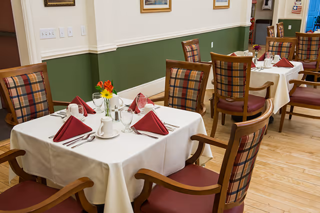 A dining room with tables covered in white tablecloths, each set with red folded napkins, glassware, and silverware. The chairs have wooden frames with plaid upholstered backs and red cushioned seats. The walls are painted green on the lower half and cream on the upper half, decorated with framed artwork.