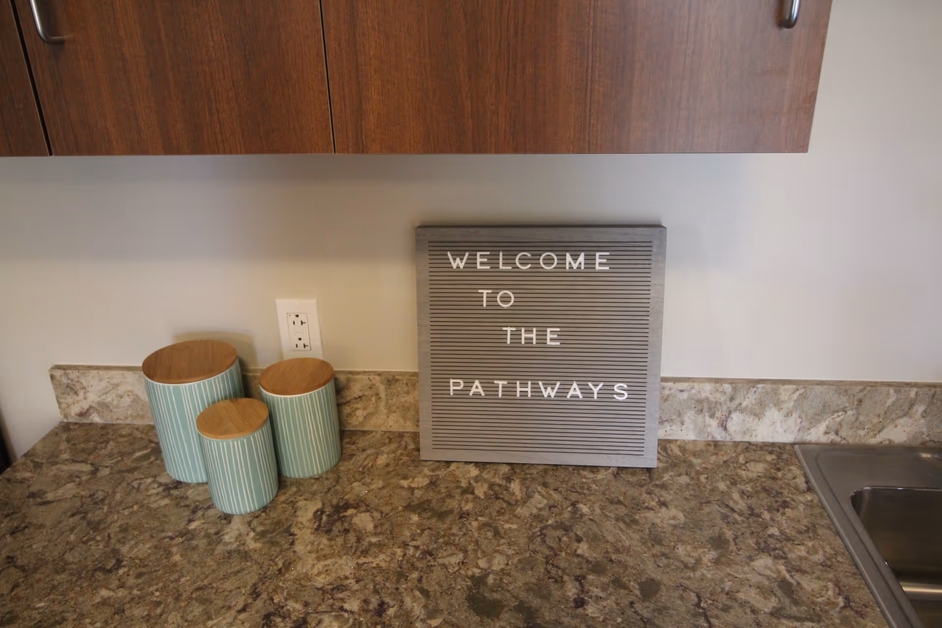 A kitchen countertop with three green and white striped canisters with wooden lids, a gray letter board with white letters spelling 'WELCOME TO THE PATHWAYS', and a stainless steel sink on the right side. Wooden cabinets are visible above the countertop.