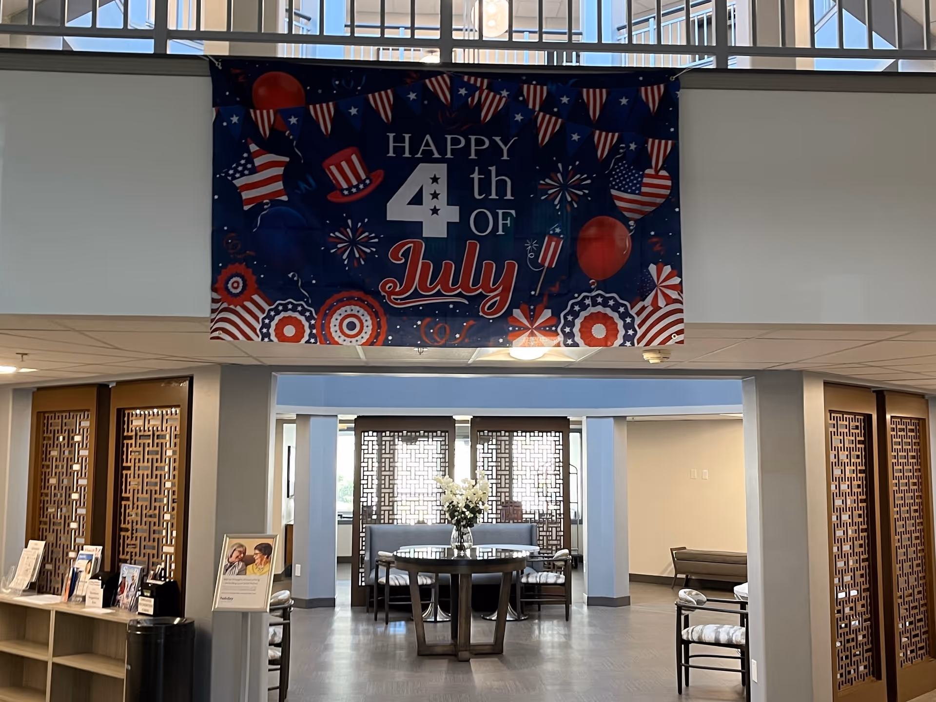 Interior view of a senior living facility lobby or common area decorated with a large 'Happy 4th of July' banner featuring patriotic designs like stars, stripes, balloons, and fireworks. The space includes a round table with a flower arrangement in the center, chairs, and decorative wooden room dividers. The area is well-lit with natural light coming through upper windows.
