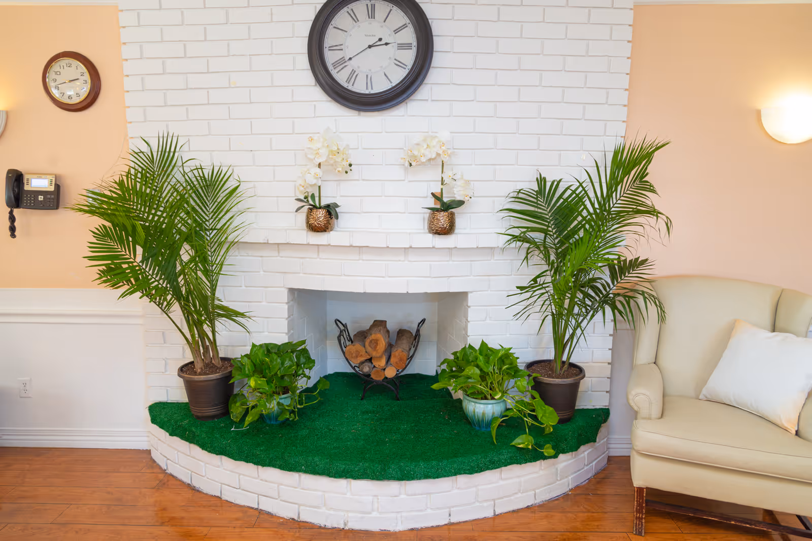 A cozy indoor corner featuring a white brick fireplace with logs inside, decorated with two potted orchids on the mantel and four green plants around the hearth. Above the fireplace is a large round clock with Roman numerals. To the right is a beige armchair with a white pillow. The walls are painted peach and white, with a wall-mounted phone and a smaller clock on the left side.