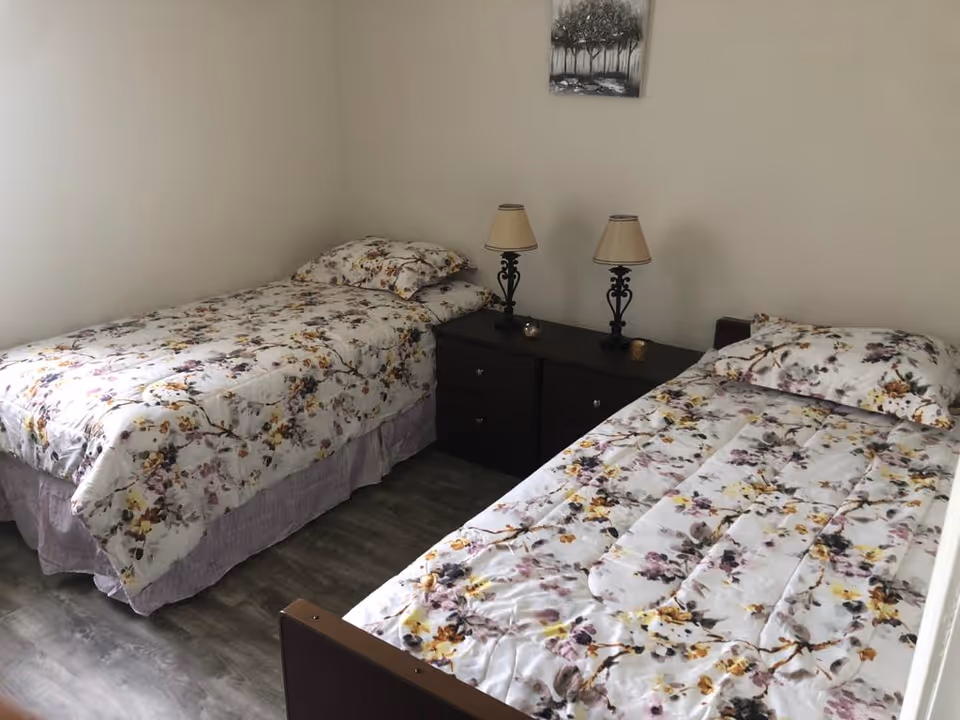 A bedroom with two single beds placed parallel to each other, each with floral patterned bedding and pillows. Between the beds is a dark wooden nightstand with two decorative lamps and small candle holders. A framed picture of trees hangs on the wall above the nightstand. The floor has a wood-like finish.