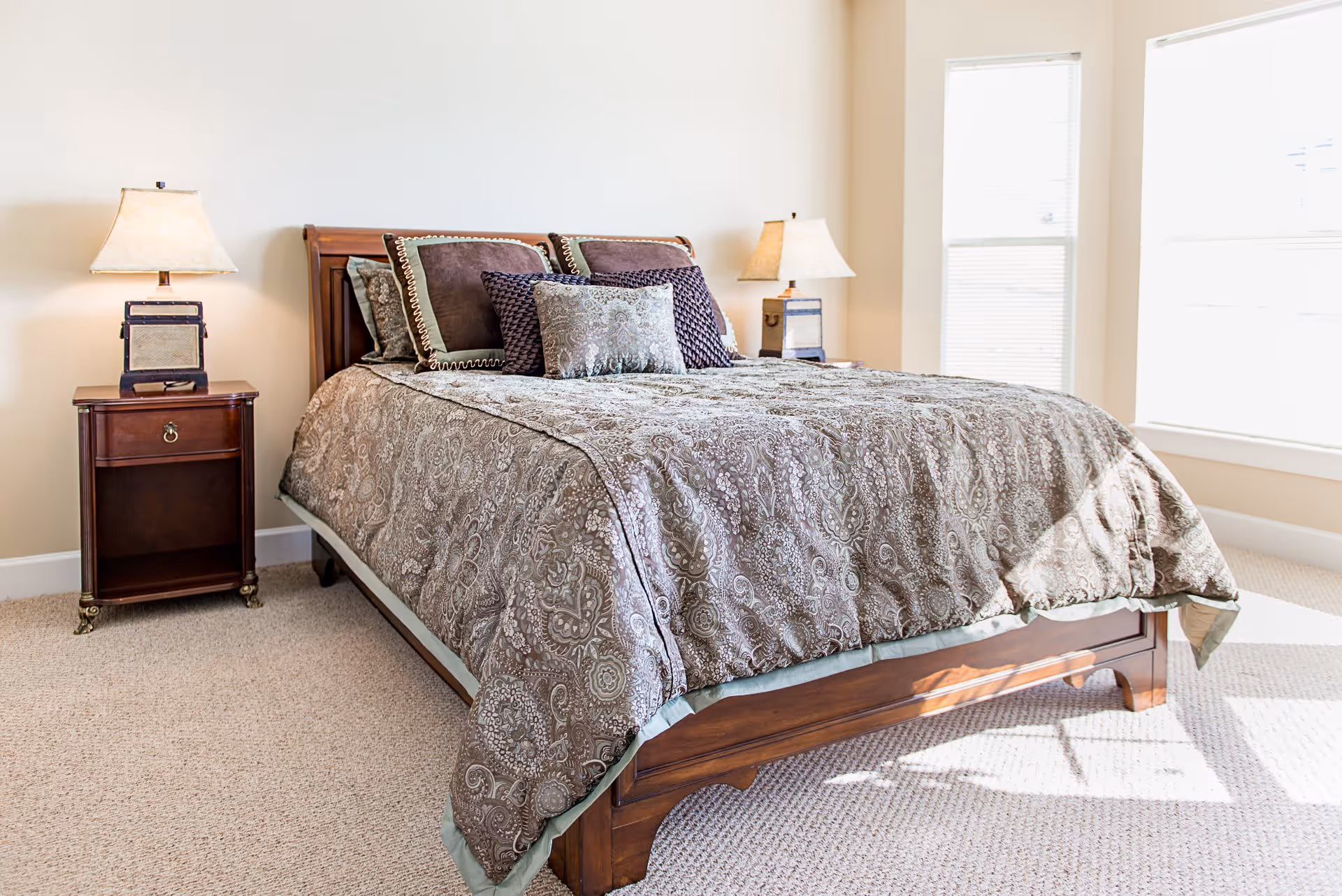 A bedroom with a wooden bed frame and a patterned brown bedspread. The bed is adorned with multiple decorative pillows in shades of brown and beige. There are two matching wooden nightstands on either side of the bed, each with a lamp featuring a beige lampshade. The room has beige walls and carpeted flooring, with two large windows allowing natural light to brighten the space.