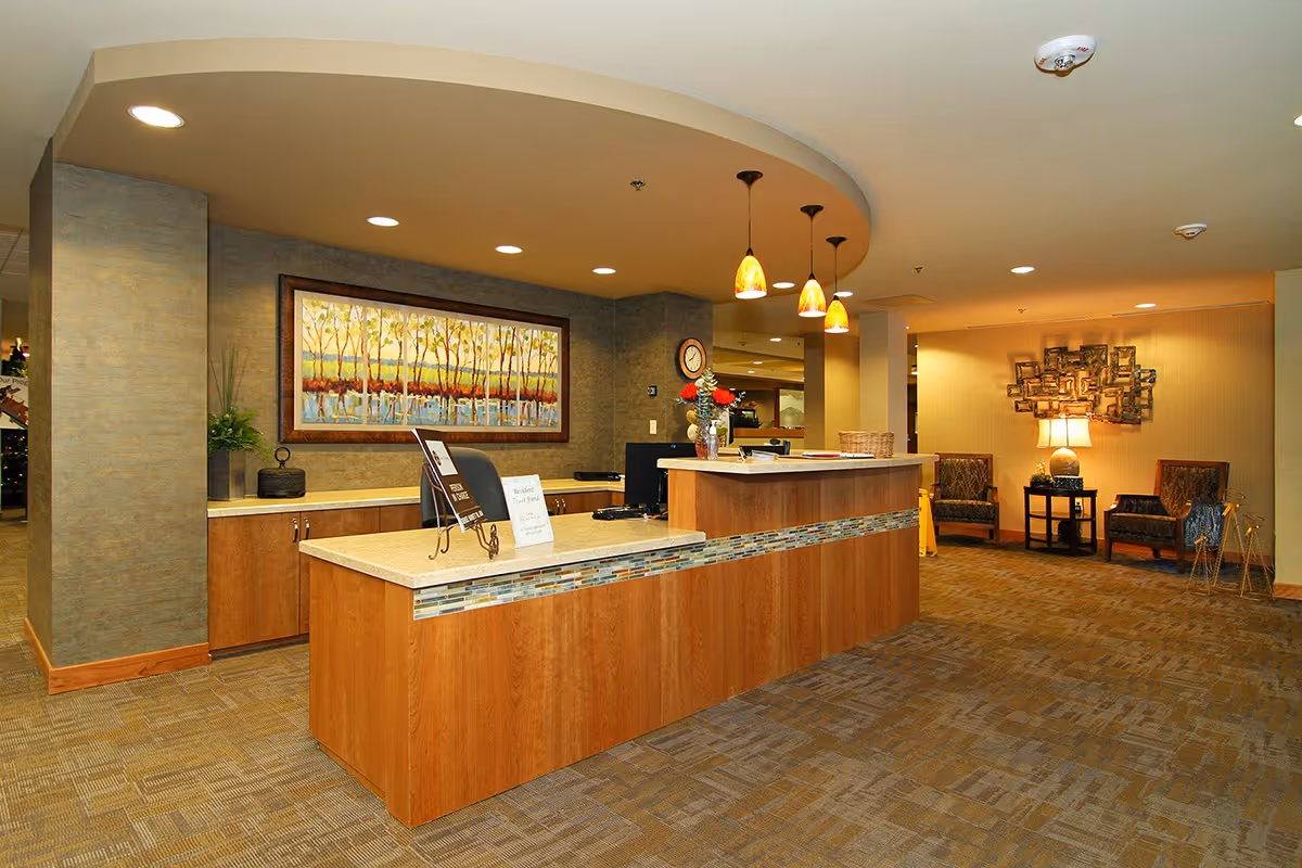 Reception area of a senior living facility with a wooden front desk featuring a decorative tile strip, pendant lights hanging from the ceiling, a large framed painting of trees on the wall behind the desk, and a seating area with two chairs, a side table with a lamp, and wall art in the background.