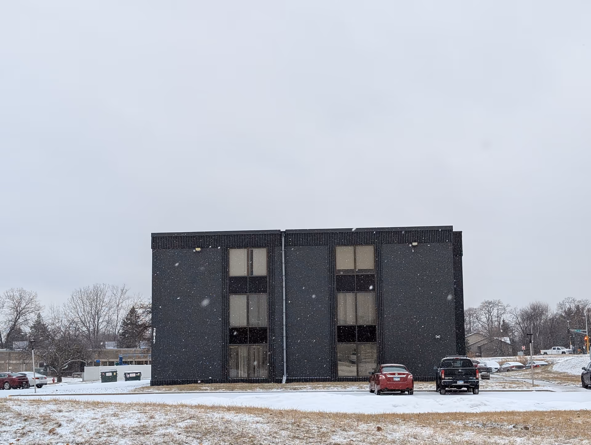 Exterior view of a dark brick three-story building with large vertical windows, surrounded by a snowy landscape and parked cars in front.