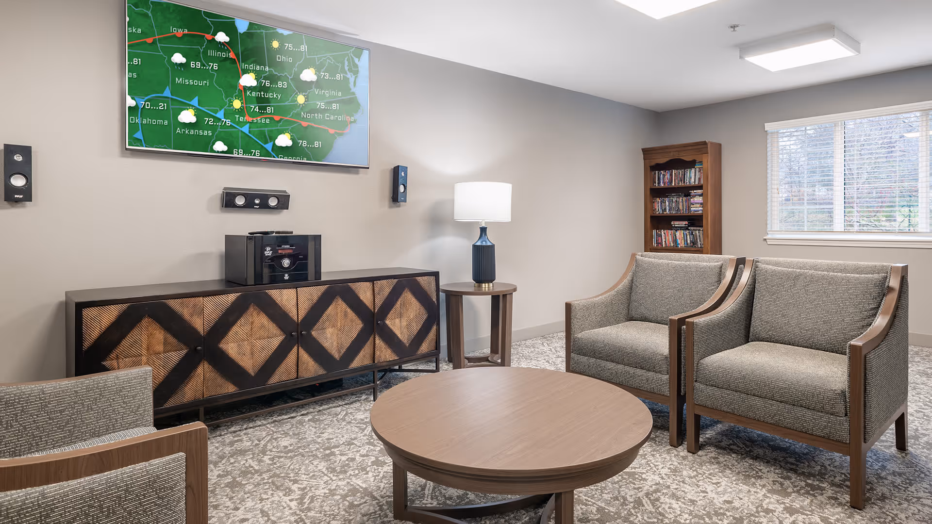 A cozy living room area with two gray upholstered armchairs and a round wooden coffee table. A wooden cabinet with a geometric pattern sits against the wall beneath a mounted flat-screen TV displaying a weather map. A small side table with a lamp is positioned between the cabinet and the chairs. A wooden bookshelf filled with DVDs or books is near a window with white blinds, allowing natural light into the room.