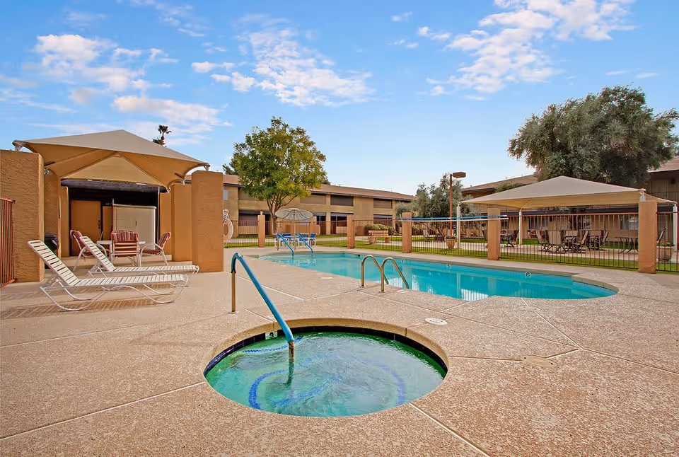 Outdoor pool area at a senior living facility with a swimming pool, a hot tub, lounge chairs, tables with umbrellas, and surrounding buildings under a partly cloudy sky.
