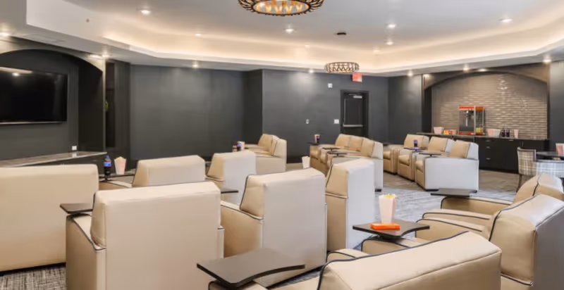 A modern lounge area with multiple beige recliner chairs arranged in rows facing a large flat-screen TV mounted on a dark gray wall. The room features soft overhead lighting, a popcorn machine on a counter in the back corner, and small tables attached to some chairs holding popcorn containers and drinks.