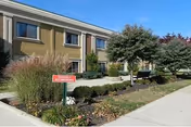 Front exterior of a two-story residential building with landscaped shrubs, trees, and a small yard sign.