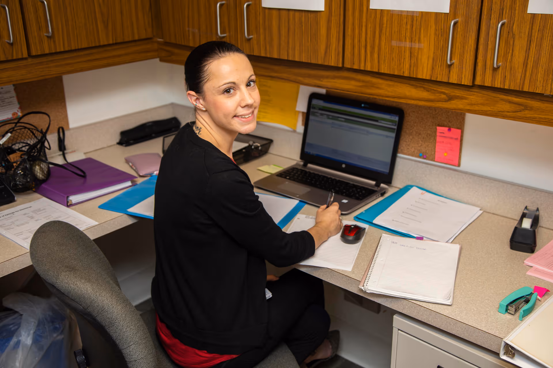 A woman sitting at a desk in an office area with wooden cabinets above. She is writing on a notepad with a laptop open in front of her, surrounded by various papers, folders, and office supplies.