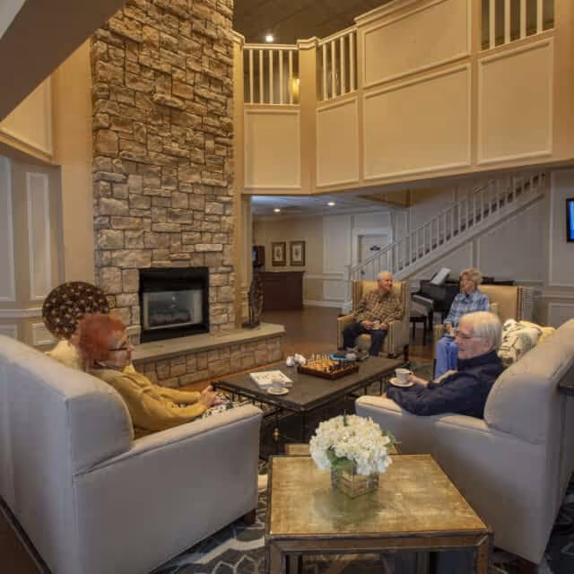 A cozy living room area in an assisted living facility with four elderly people sitting on sofas and armchairs around a coffee table. The room features a tall stone fireplace, a piano in the background, and a staircase leading to an upper floor. A small table with a flower arrangement is in the foreground.
