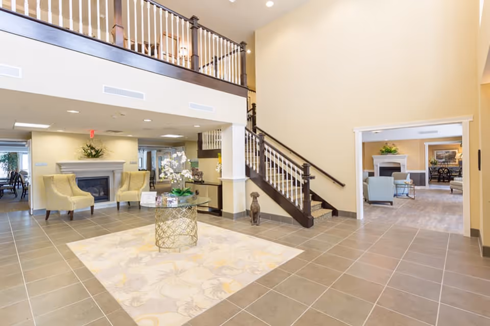Bright two-story lobby with a decorative round table on a rug, a staircase, seating areas and a fireplace.