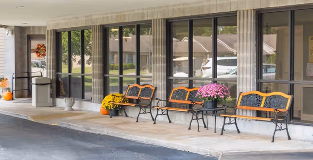 Outdoor covered seating area with three black and wood benches and a small table holding a pot of pink flowers. There are pumpkins and yellow flowers near the benches, large windows reflecting nearby buildings and vehicles, and a trash can near a door decorated with a fall wreath.