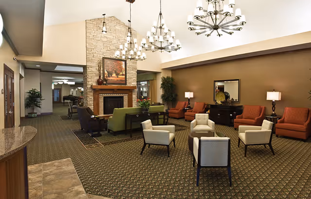 Spacious senior living lounge with grouped chairs and sofas around a stone fireplace, chandeliers, and decorative plants.