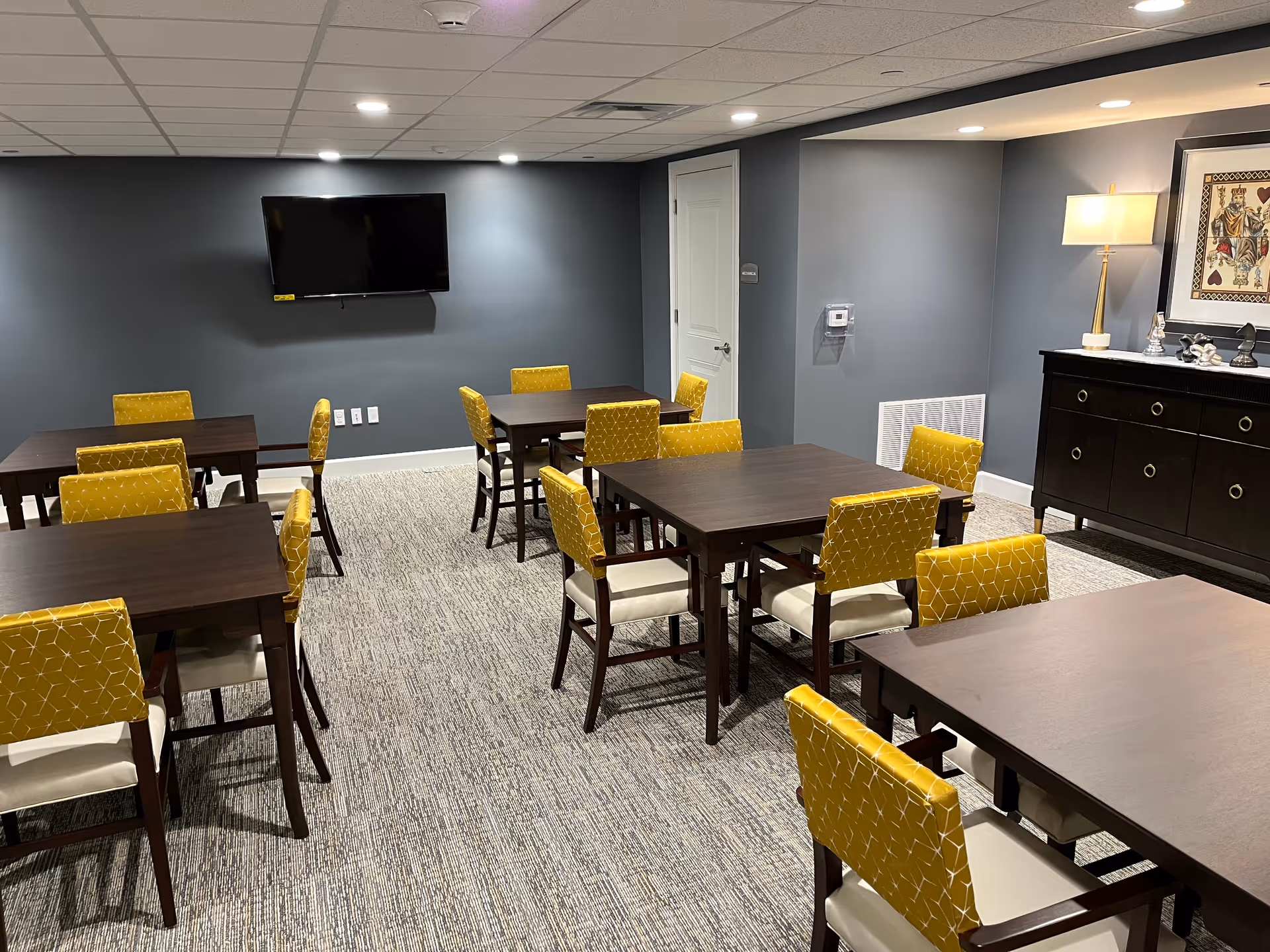Interior room with multiple dark wood tables and yellow cushioned chairs arranged neatly on a patterned carpet. A flat-screen TV is mounted on a gray wall, and a dark wood sideboard with a lamp and framed artwork is visible on the right side.