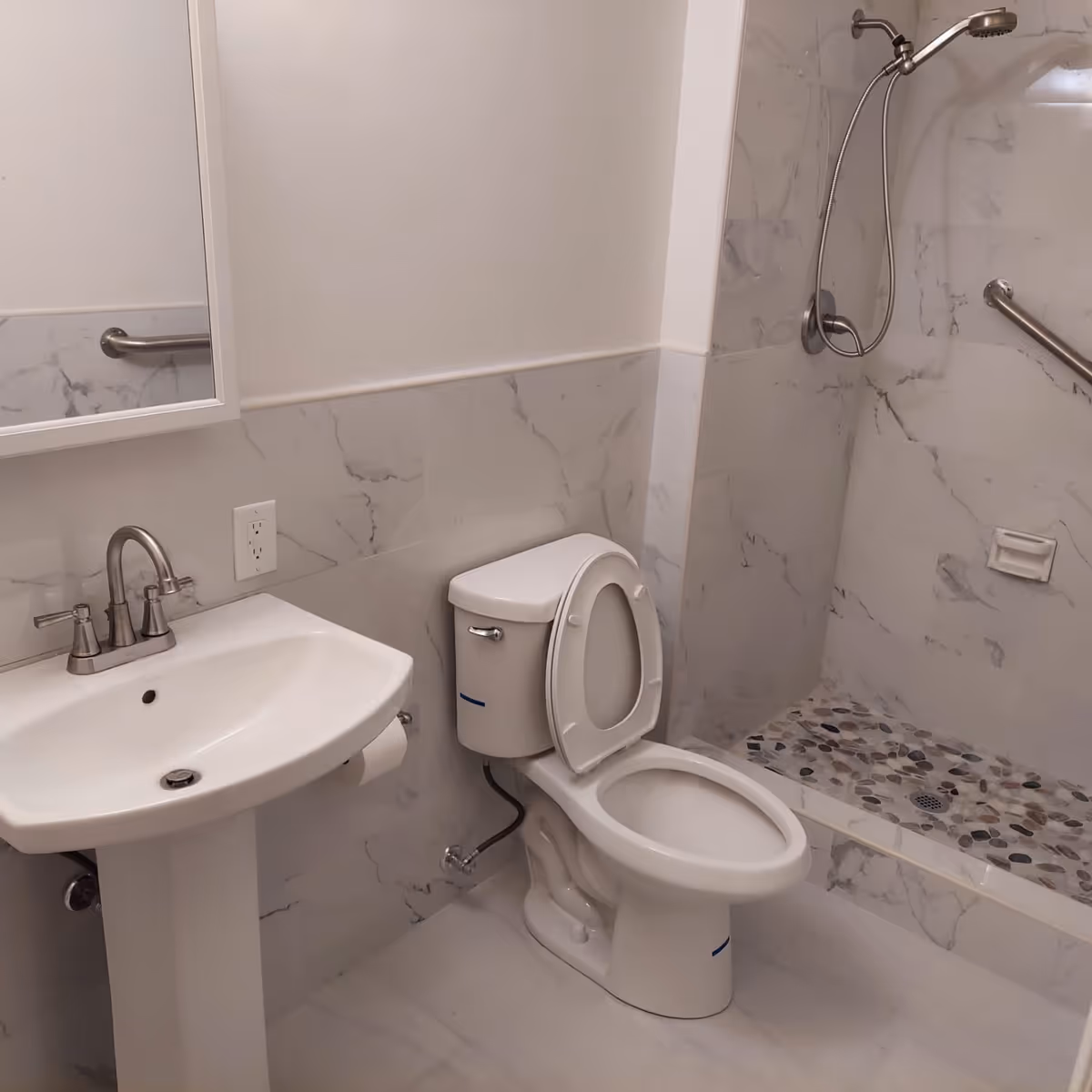 A clean bathroom with a white pedestal sink, a toilet with the lid open, and a walk-in shower with a handheld showerhead and grab bars. The walls and floor have a marble-like tile design.