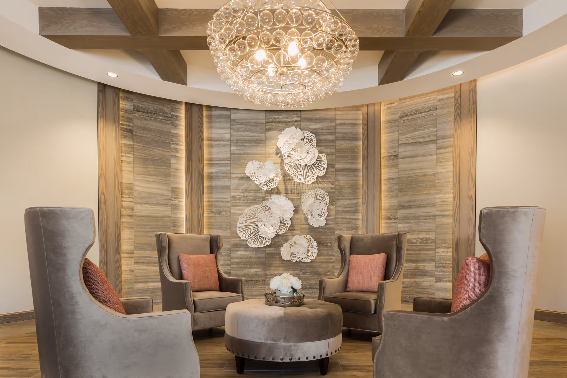 A cozy seating area with four high-back armchairs arranged around a round ottoman. Each chair has a coral-colored cushion. The background features a textured wall with decorative white floral wall art. Above, a large, elegant chandelier with multiple glass spheres hangs from a ceiling with wooden beams.