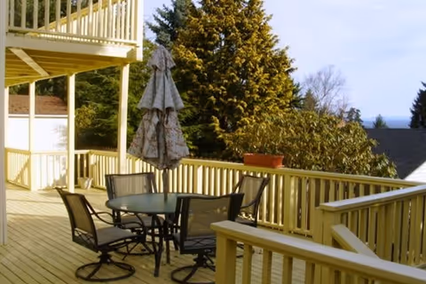 Wooden outdoor deck with a round glass table, four chairs and a closed patio umbrella overlooking trees and neighboring rooftops.