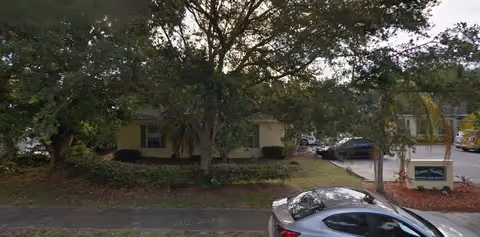 Exterior view of Spring Hills Assisted Living Facility partially obscured by large trees and landscaping, with a parking lot visible to the right and a silver car parked in front.