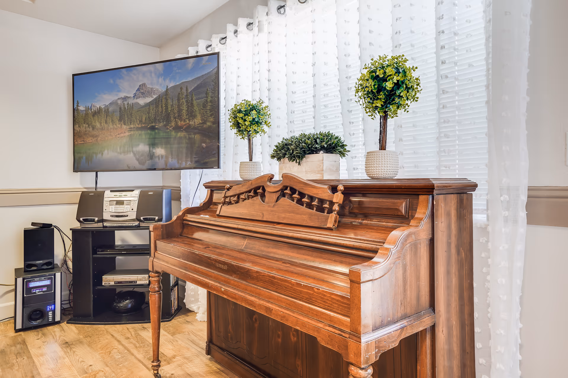 A wooden upright piano with decorative carved details is placed in front of a window with sheer white curtains. On top of the piano are three potted green plants. To the left of the piano, there is a black entertainment stand holding a stereo system, speakers, and other electronic devices. A flat-screen TV mounted on the wall displays a scenic landscape of mountains, trees, and a lake.