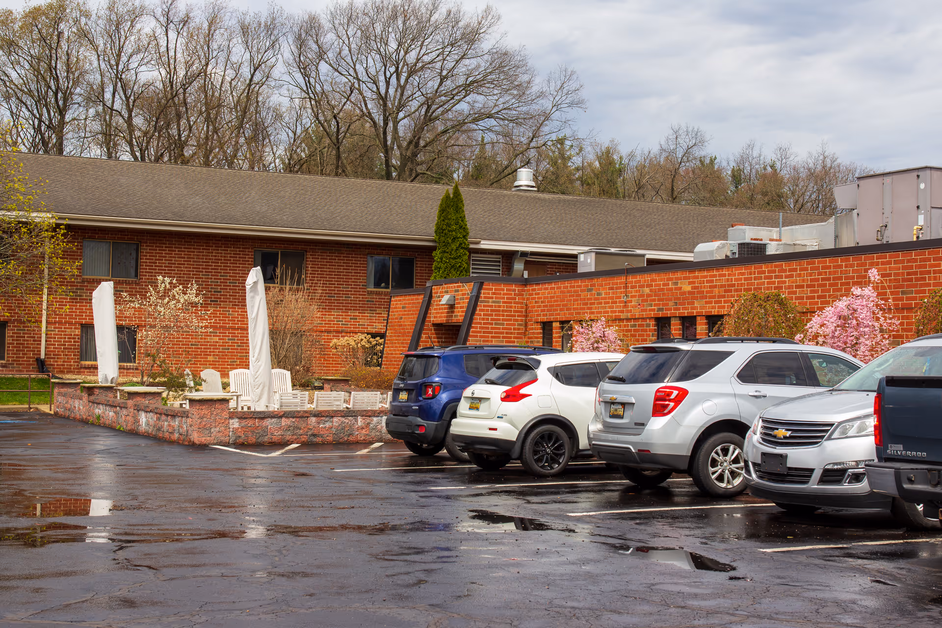 Parking lot with several parked cars in front of a brick building with outdoor seating area and closed umbrellas, surrounded by trees and some flowering bushes under a cloudy sky.