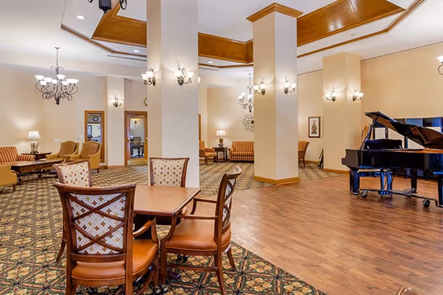 A spacious senior living facility common area with patterned carpet and wood flooring. There are several wooden chairs with cushioned seats around a small table in the foreground. In the background, there are sofas and armchairs arranged for seating, wall sconces providing lighting, and a grand piano on the wood floor section. The ceiling has recessed lighting and decorative molding.