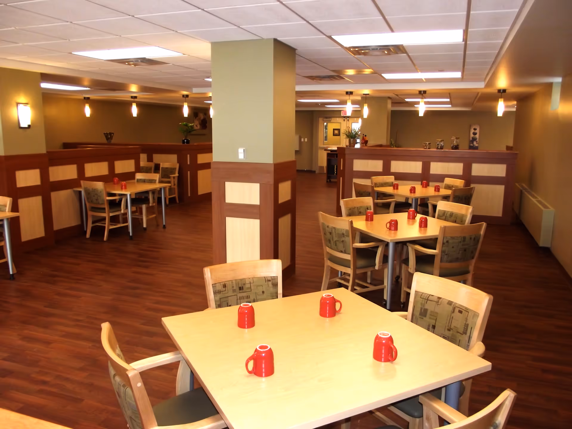 A dining room in a senior living facility with multiple wooden tables and chairs arranged neatly. Each table has red mugs placed upside down. The room has wood flooring, green walls with wooden paneling, and ceiling lights providing bright illumination. There are decorative plants and wall art in the background.