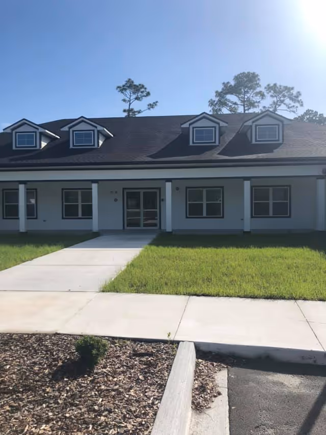 Front exterior view of a single-story building with a dark roof and white walls, featuring four dormer windows and a central entrance with double glass doors. A concrete walkway leads up to the entrance, surrounded by green grass and a mulched area with a small plant in the foreground. Tall pine trees are visible in the background under a clear blue sky.