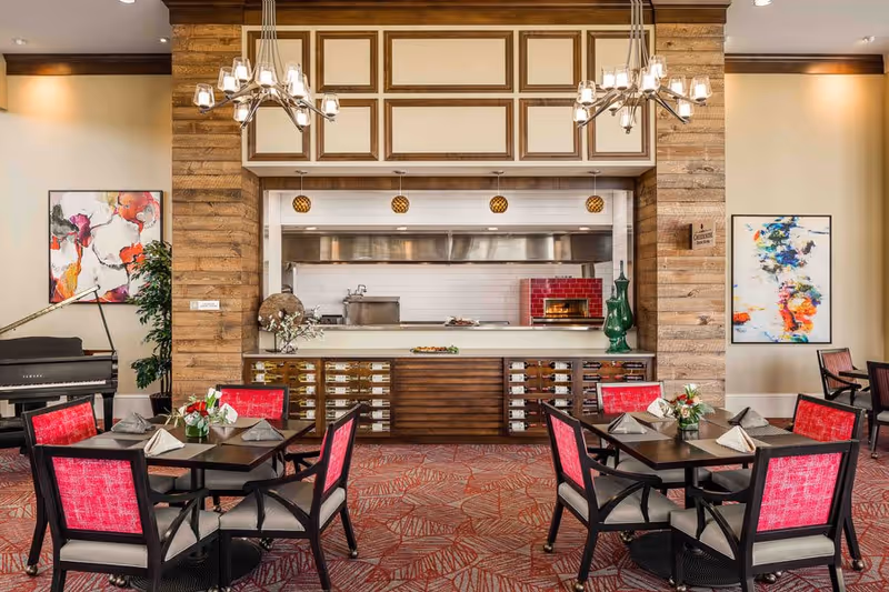 A dining area with two square tables, each surrounded by four chairs with red upholstery. The tables are set with napkins and small floral centerpieces. Behind the tables is an open kitchen with a wood-paneled wall, white cabinetry, and a red brick pizza oven. Two modern chandeliers hang from the ceiling. Colorful abstract paintings and a black grand piano are visible on the side walls.