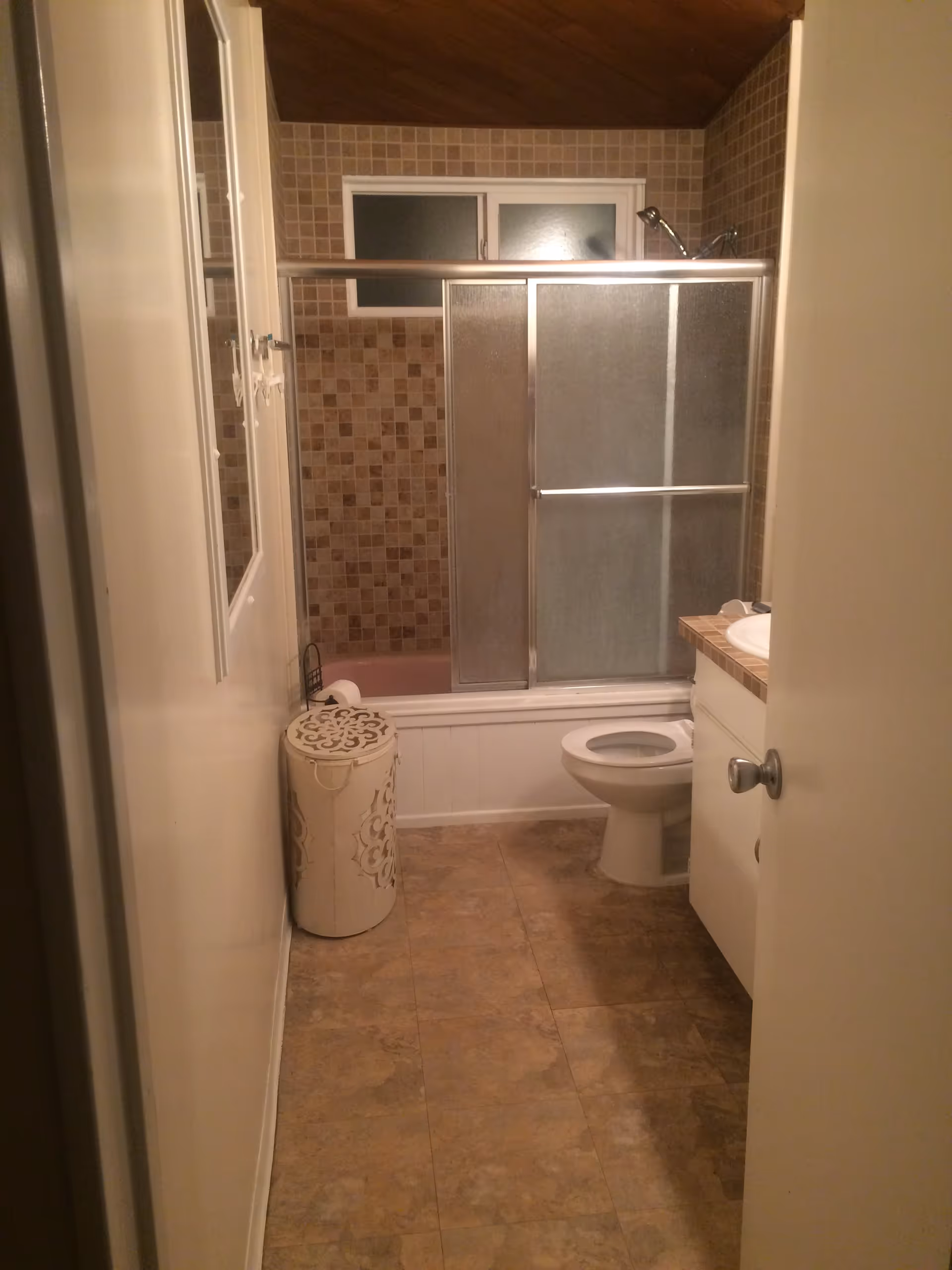A small bathroom with a tiled floor and walls. There is a bathtub with sliding glass doors and a showerhead, a toilet, a white cabinet with a sink, and a decorative white laundry hamper. The ceiling is wooden and slanted, and there is a frosted window above the bathtub.