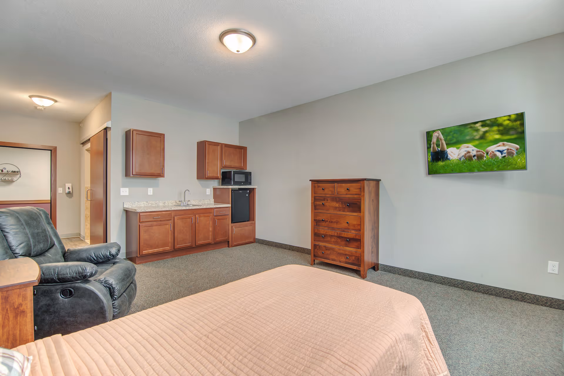 A furnished senior living bedroom with a bed in the foreground, a recliner, a small kitchenette with cabinets and sink, a wooden dresser, and a wall-mounted TV.