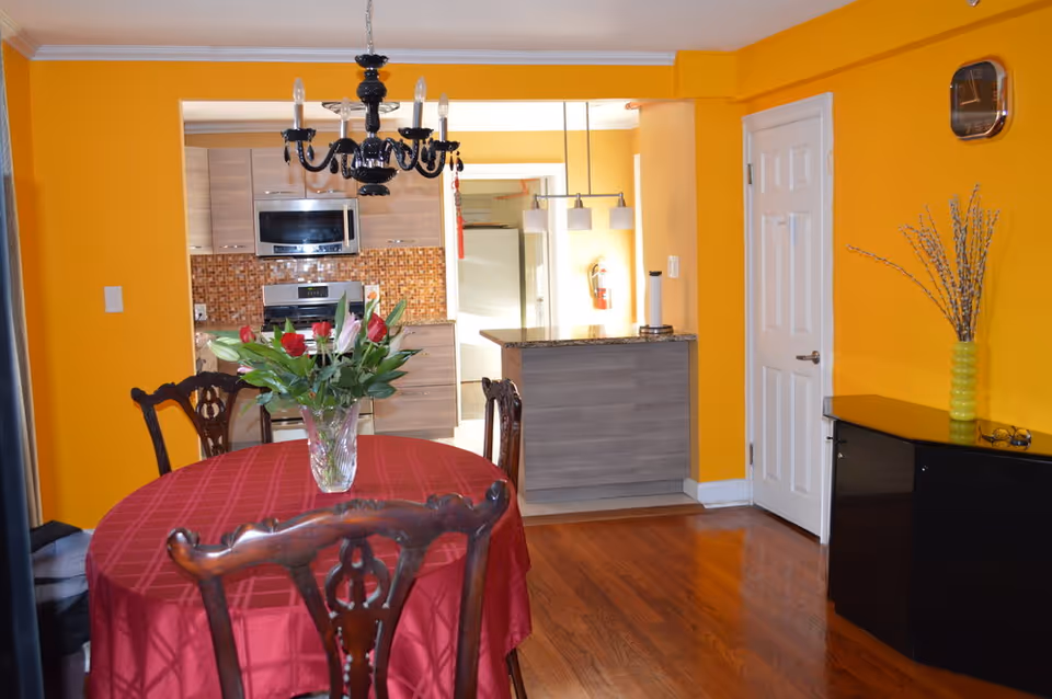 A dining area with a round table covered with a red tablecloth and a vase of flowers in the center. The room has bright yellow walls and wooden flooring. There are four wooden chairs around the table. In the background, there is a kitchen with modern cabinets, a microwave, and a stove. A black chandelier hangs above the dining table. To the right, there is a black cabinet with a green vase and decorative branches, and a wall clock above it.
