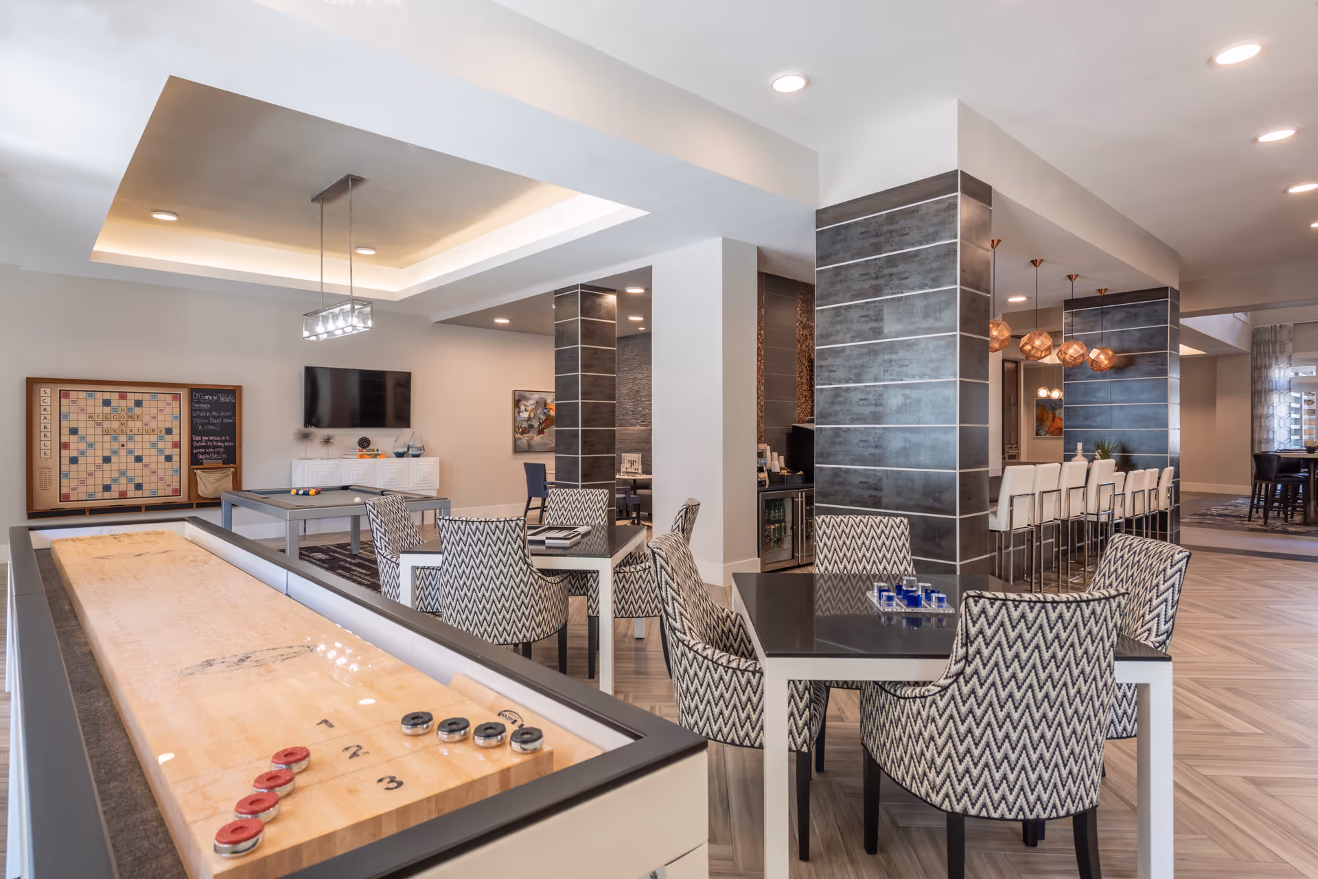 A modern recreational room with a shuffleboard table in the foreground, several tables with patterned chairs, a pool table, a large Scrabble board on the wall, a flat-screen TV, and a bar area with high chairs and pendant lighting in the background.