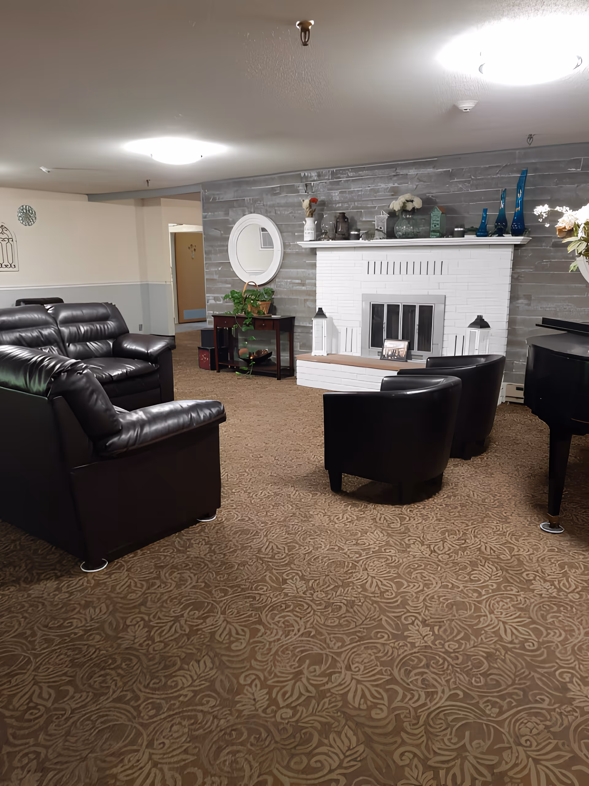 A cozy living room area with a patterned carpet, black leather sofas and chairs, a white brick fireplace with decorative items on the mantel, a round mirror on the wall, and a small table with a plant underneath the mirror.