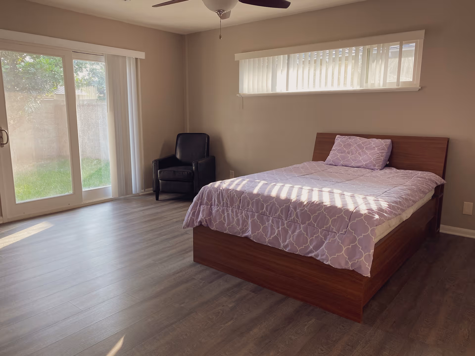 A bedroom with a wooden bed frame and a lavender patterned comforter and pillow. There is a black armchair in the corner near sliding glass doors that lead outside to a grassy area. The room has light-colored walls, a horizontal window with vertical blinds, and wood flooring.