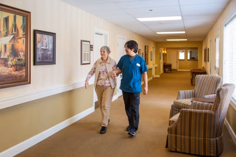 An elderly woman walking down a hallway in an assisted living facility, accompanied by a caregiver in blue scrubs. The hallway is decorated with framed paintings and has comfortable armchairs along the wall with large windows letting in natural light.