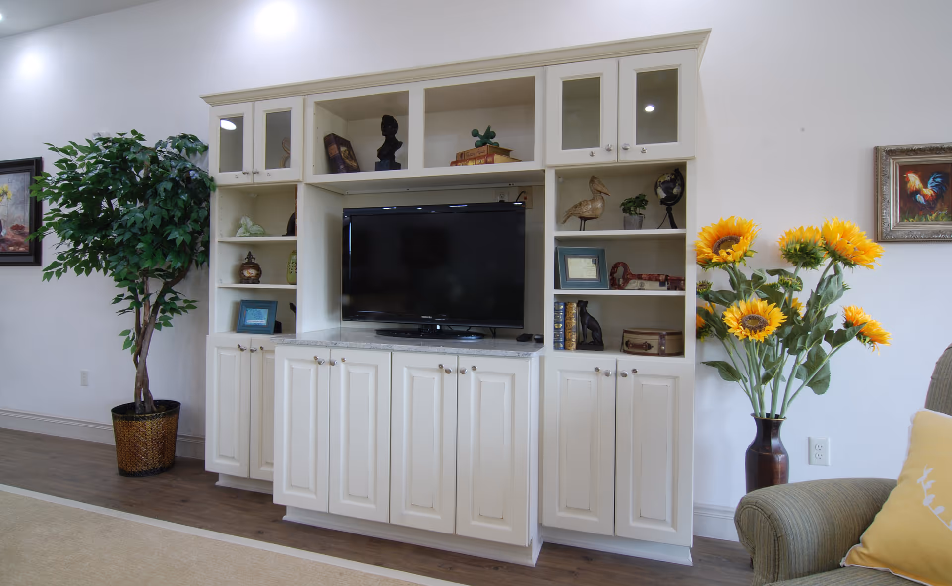 A cozy living room area featuring a white entertainment center with a flat-screen TV, decorative shelves with books, sculptures, and framed pictures. To the left is a tall potted plant, and to the right is a vase with bright yellow sunflowers next to a green armchair with a yellow pillow. A framed painting of a rooster hangs on the wall.