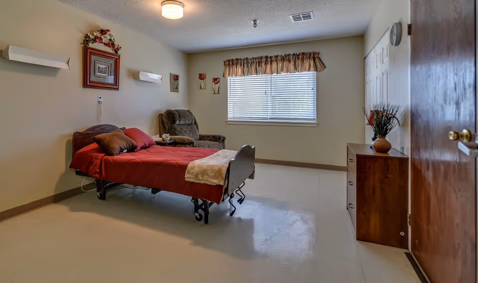 A simple nursing center bedroom with a single bed covered in a red blanket and brown pillows. There is a cushioned armchair next to the bed, a wooden dresser with a vase of flowers, and a window with blinds and a valance. The walls are decorated with framed pictures and floral artwork.
