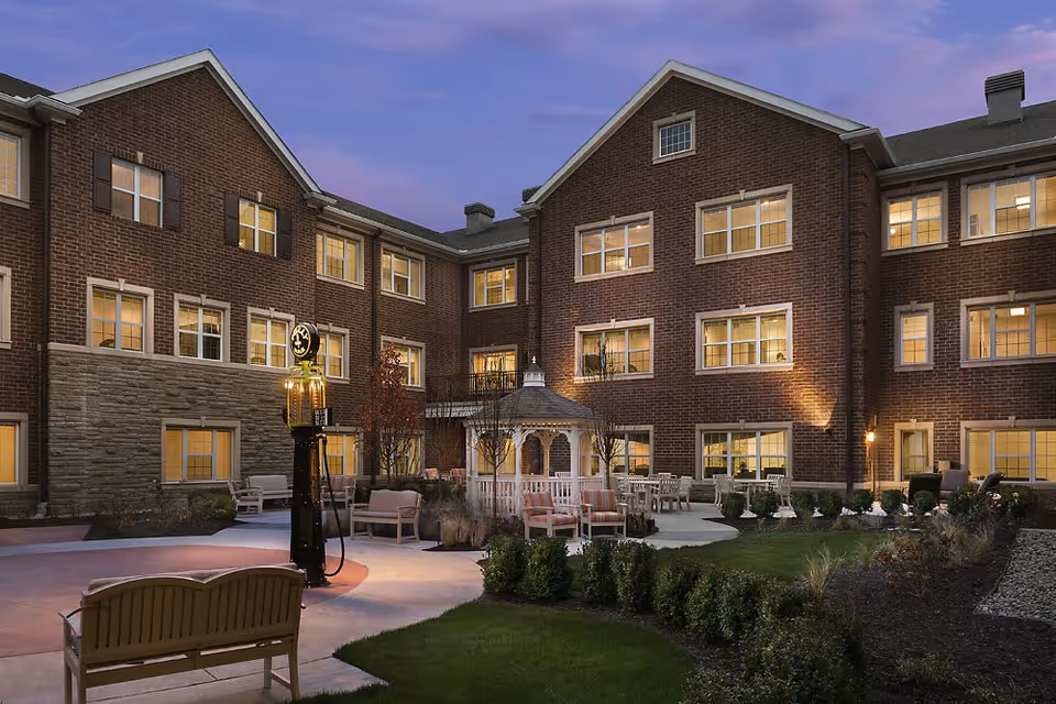 Evening view of a senior living facility courtyard with a brick building surrounding it. The courtyard features a white gazebo, outdoor seating with benches and chairs, well-maintained landscaping, and a vintage-style lamp post. The building windows are lit from inside, creating a warm and inviting atmosphere.