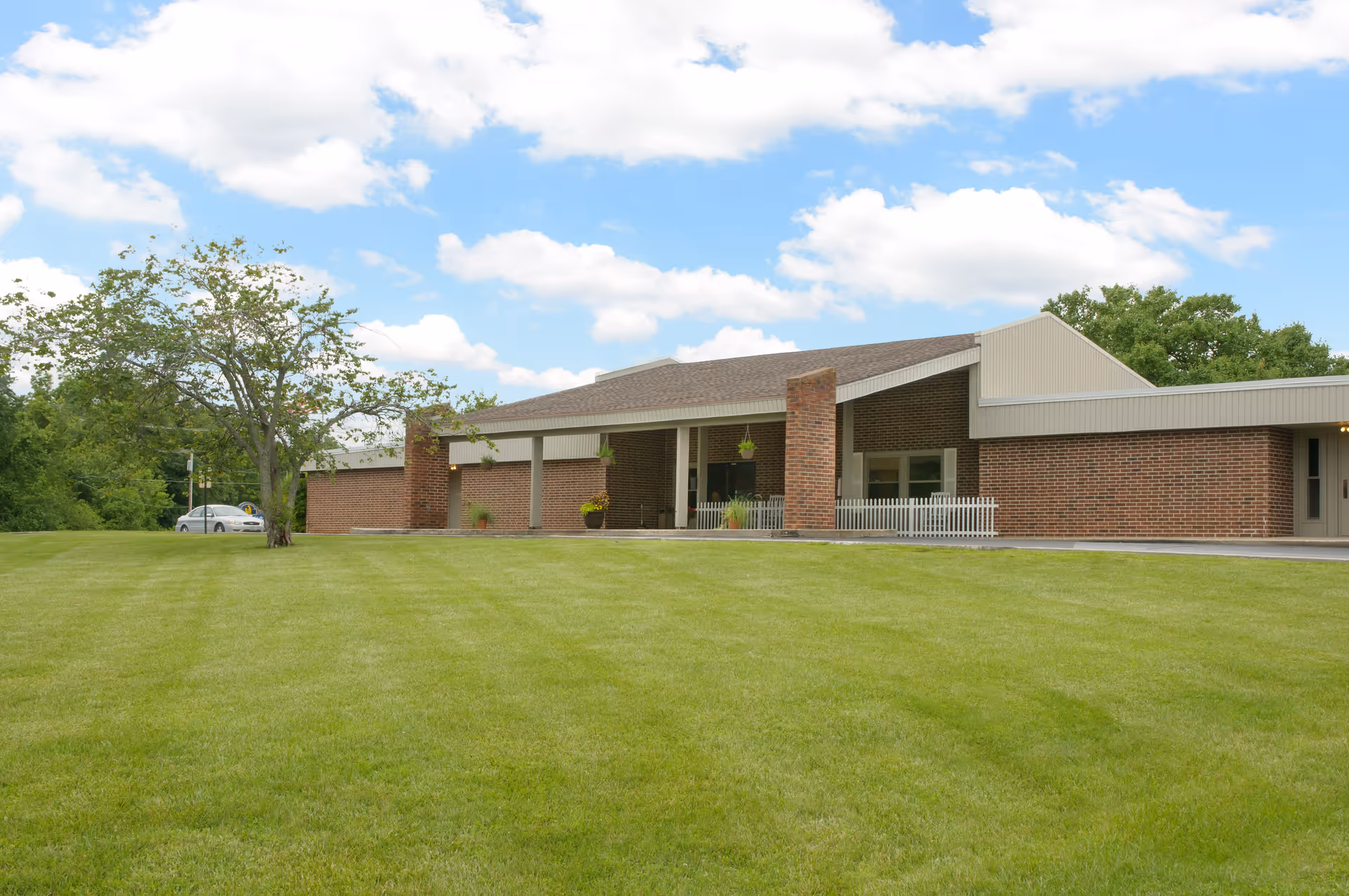 Single-story brick building with a covered entrance facing a wide green lawn and a tree under a partly cloudy sky.