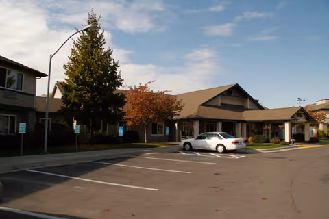 Exterior view of a senior living facility building with a parking lot in front. There are a few trees, including one with autumn-colored leaves, and a white car parked near the entrance. The sky is partly cloudy.