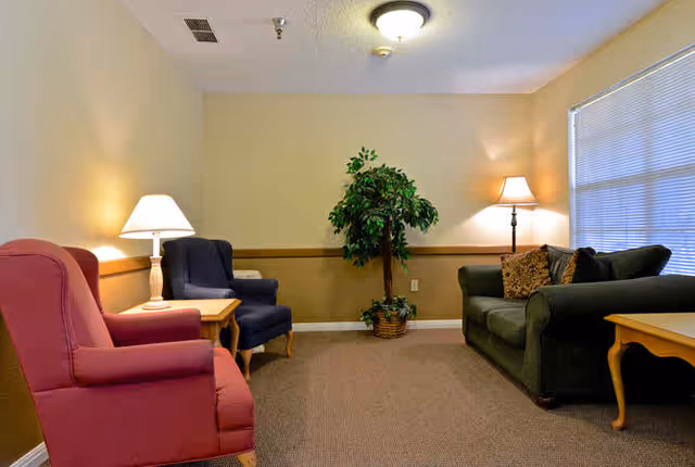 A cozy living room with a red armchair, a blue armchair, and a dark green sofa arranged around two wooden side tables. There are two lamps providing warm lighting and a potted plant in the corner. A large window with blinds is on the right wall.