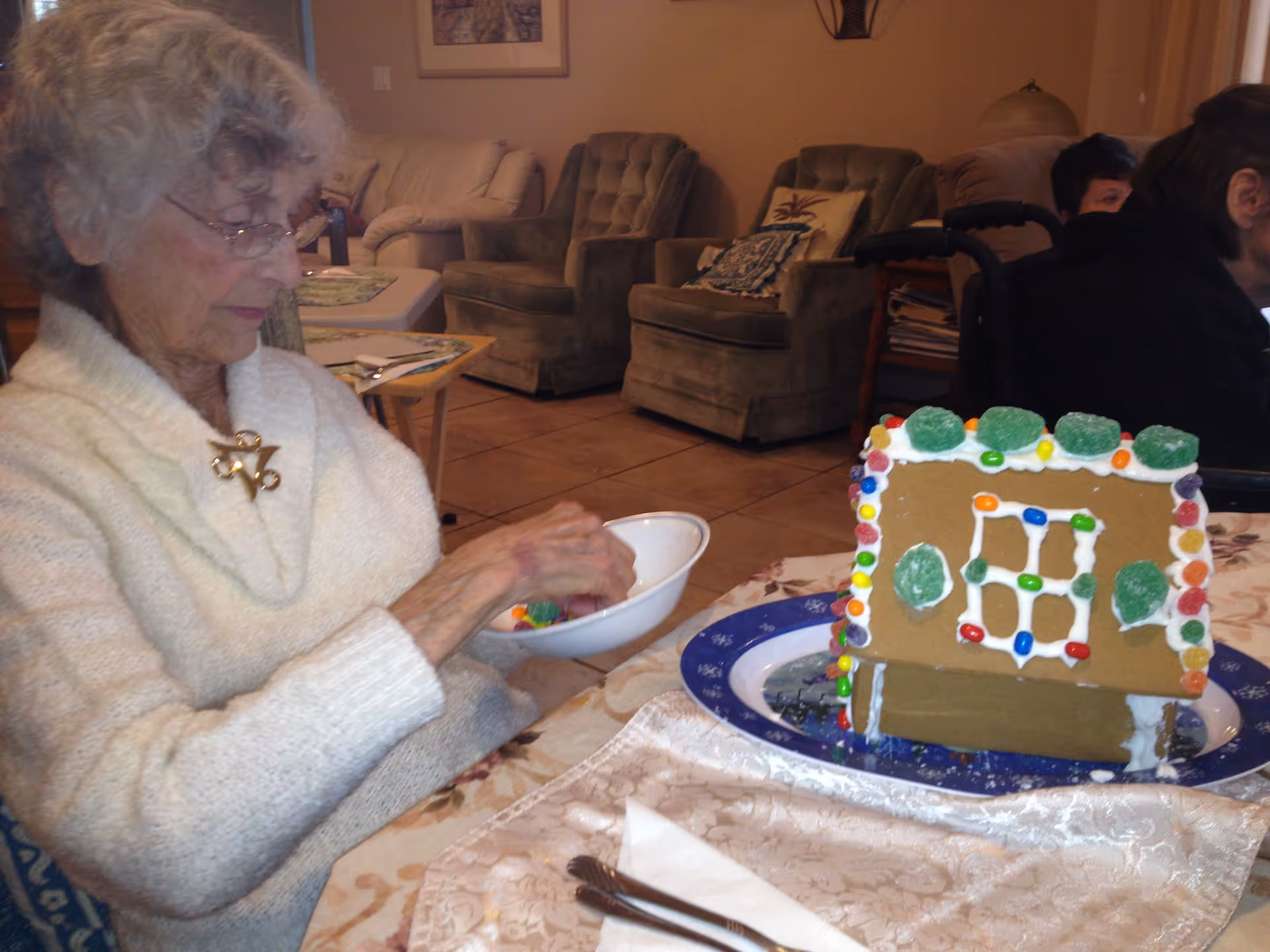 An elderly woman wearing glasses and a white sweater is sitting at a table in a living room setting. She is holding a white bowl with colorful candies and appears to be decorating a gingerbread house placed on a blue and white plate in front of her. The background shows cushioned chairs, a couch, and another person partially visible.